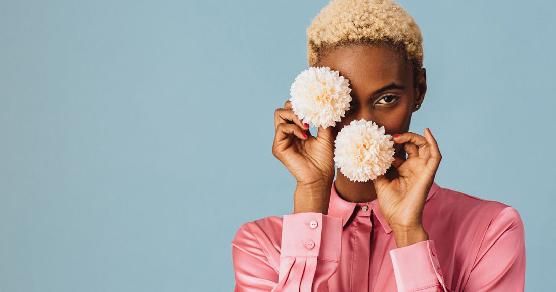 A black woman with short hair in a pink shirt holding two flowers in front of her face.